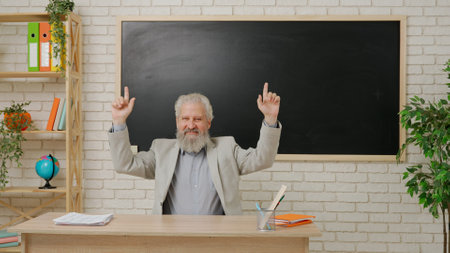 Aged man college professor sit at desk in classroom in front of chalkboard pointing at empty space holding hands up. Education concept.の写真素材