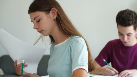 High school teenagers in classroom, girl sit at desk writing test, looks at camera smiles and shows thumb up. Modern education concept.の写真素材