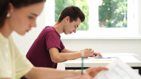 High school teenagers in classroom, girl sit at desk writing test, looks at camera smiles and shows thumb up. Modern education concept.の写真素材