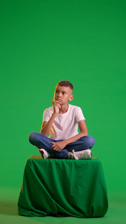 Pensive boy sitting on a stool on a green screenの写真素材