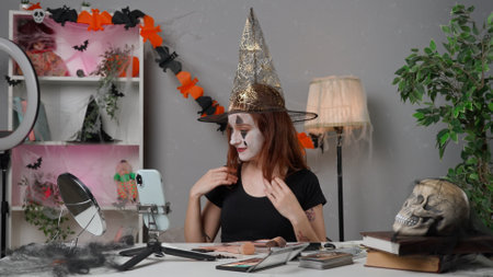 Young woman beauty blogger at home studio films video tutorial for halloween party makeup on smartphone, wears spooky witch hat and talks with followers.の写真素材