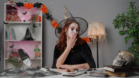 Young woman beauty blogger in scary cape at home studio films video tutorial for halloween party makeup, talks at camera, wears posh witch hat and smiling.の写真素材