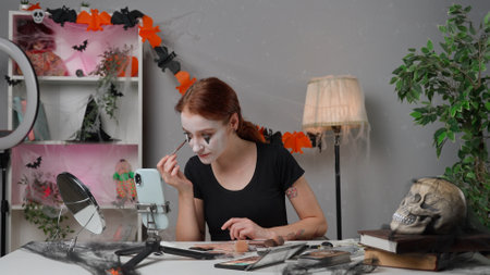 Young woman beauty blogger at home studio films video tutorial for halloween party makeup on smartphone, applies black cream paint under eye for scary look.の写真素材