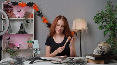 Young woman beauty blogger at home studio films video tutorial for halloween party makeup on smartphone, greeting followers showing cosmetic products.の写真素材