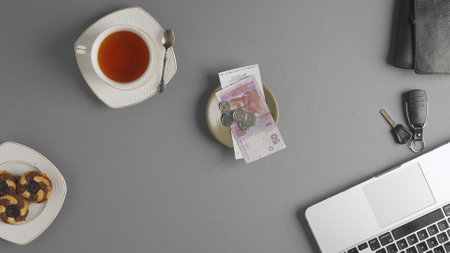 Top view of a saucer with a check for payment, coins and hryvnia bills, next to a cup of tea and a laptop, wallet and car keys on the table. The concept of money and payment for services.の写真素材