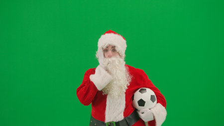 Portrait of man dressed as Santa on chroma key green screen background. Santa Claus in red outfit holding football ball and cheering at camera.の写真素材