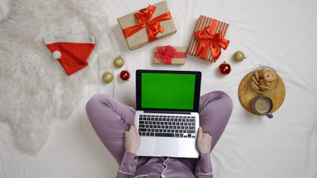 Top view female on the bed using gadget chroma key green screen. Girl in pajama on the bed with gifts and snacks holding laptop.の写真素材