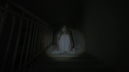 Creepy ghost woman in white dress and scary makeup alone on staircase in dark haunted house lighted by flashlight. Halloween horror advertisement concept.の写真素材