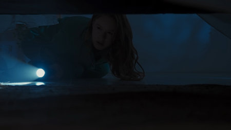Woman on the floor in dark room checking space under bed with flashlight, looking around for...