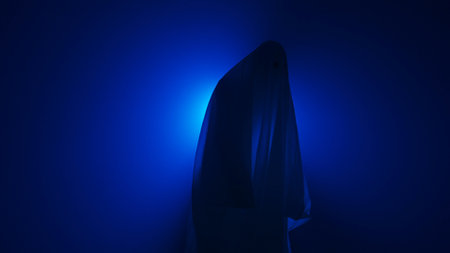 Funny Halloween ghost in white sheet makes scary gestures on background with blue spotlight in...