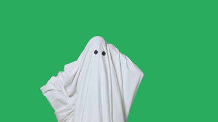 Funny halloween ghost in white sheet confused thinking, raises hand with idea, isolated on chroma key green screen background. Happy halloween spooky concept.の写真素材