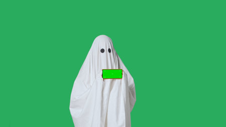 Funny halloween ghost in white sheet pointing shows thumb up at smartphone isolated on chroma key...