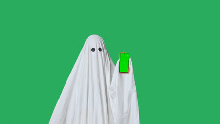 Funny halloween ghost in white sheet pointing shows thumb up at smartphone isolated on chroma key...