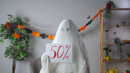 Funny halloween ghost in white sheet with black eyes in bright room holding showing paper sign with...