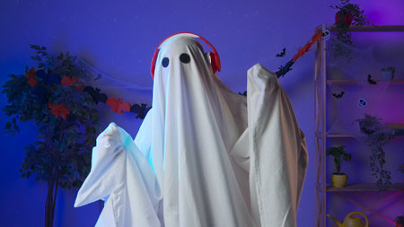 Funny halloween ghost in white sheet with black eyes in dark room dancing and listening music in...