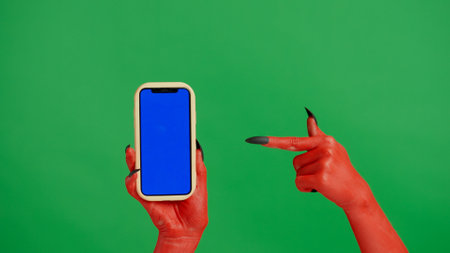 Creepy halloween devil monster hand red paint with black nails holds smartphone points finger,...