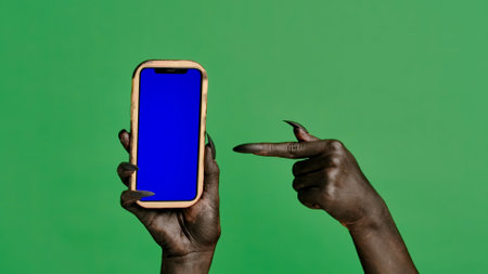 Creepy halloween devil monster hand black paint with nails holds smartphone points finger, isolated on chroma key green screen background, workspace mockup.の写真素材