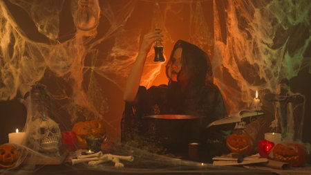 A witch in black clothes is cooking a potion from a spell book in a scary kitchen with pumpkins and...