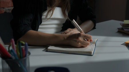 Woman sitting at desk writing notes in dairy notebook with pen, concentrated person writing down ideas and thoughts, close up shot.の写真素材
