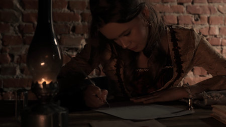 Antique young woman in renaissance dress sitting writing letter with vintage quill pen with ink on...