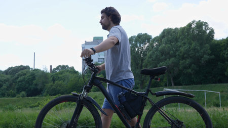 A man walking in a park with a bicycle in his hands. A cyclist walking in a green park on a sunny...