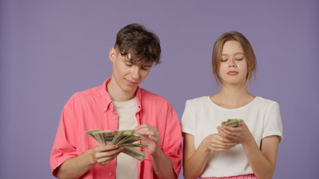 Positive couple of young boyfriend and girlfriend in pink white clothes isolated on purple background waving fan of money bills, spilling cash having fun smiling.の写真素材