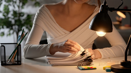 Workaholic woman sitting at office desk searching through overload stack of paper documents with...