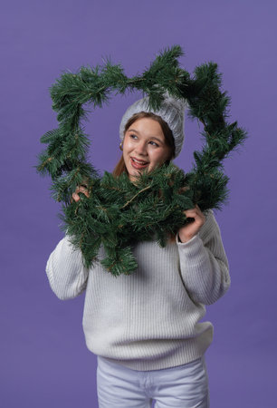 Cheerful young woman in white clothing and knitted hat posing with christmas wreath, isolated on purple studio background.の写真素材
