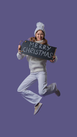 Cheerful young woman in white clothing and knitted hat jumps holding chalkboard with merry christmas text, isolated on purple studio background. Full body.の写真素材