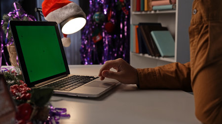 Adult business man sitting at desk working late at night in christmas decorated office, using laptop with chroma key green screen background, workspace mockup.の写真素材