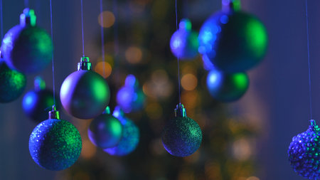 Christmas tree balls in green blue neon light, sparkling colorful toys hanging from above, sparkling ornaments garland close up shot on blurred festive background.の写真素材