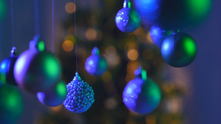 Christmas tree balls in green blue neon light, sparkling colorful toys hanging from above, sparkling ornaments garland close up shot on blurred festive background.の写真素材