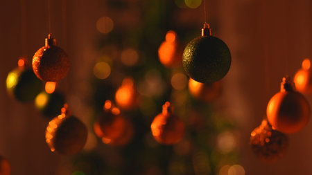 Christmas tree balls in red orange warm light, sparkling colorful toys hanging from top, ornaments garland close up shot on blurred festive background.の写真素材