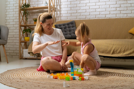 Young mother and daughter sitting on the floor at home spending time together, playing and laughing, middle shot.の写真素材