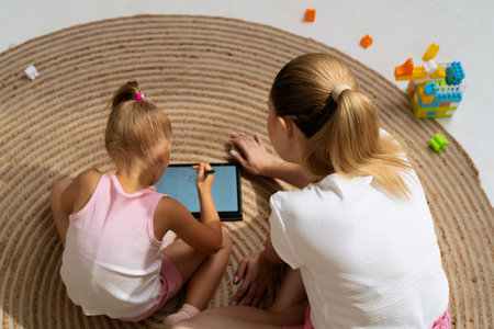 Young mother and daughter sitting on the floor at home spending time together, girl drawing with stylus on tablet mom helping, top view.の写真素材