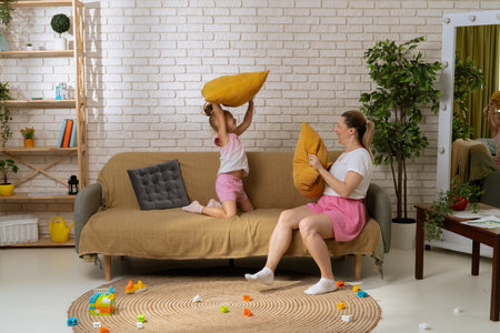 Young mother and daughter sitting on sofa at home spending time together, playing pillow fight, laughing and smiling, full shot.の写真素材