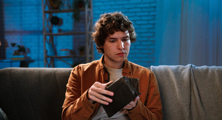 Young man spending evening at home sitting on sofa in living room, holding wallet and looking inside with serious focused face expression.の写真素材