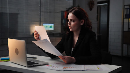 Young business woman sits at desk works late night in office, using laptop, reading documents, concentrated focused expression, side shot.の写真素材