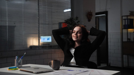 Young business woman sits at desk works late night in office, using laptop, sits back on chair hands...