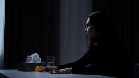Silhouette of woman suffering mental health issues, sitting at the table in front of camera, depressed expression, scatters pills on the table, side shot.の写真素材