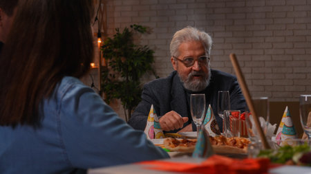 Family sit at table at home, grandchildren and parents celebrating birthday of grandfather, family members enjoying meal, talking and smiling.の写真素材