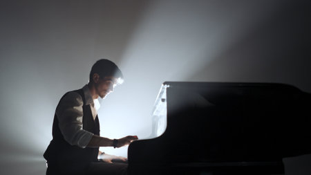 Pianist sitting and playing on the black grand piano on dark concert stage against bright spotlight, isolated on black studio background. Artist performing in concert hall.の写真素材