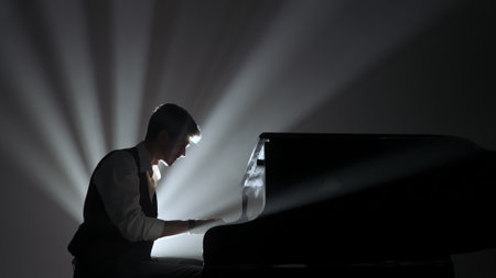 Pianist sitting and playing on the black grand piano on dark concert stage against bright spotlight, isolated on black studio background. Artist performing in concert hall.の写真素材