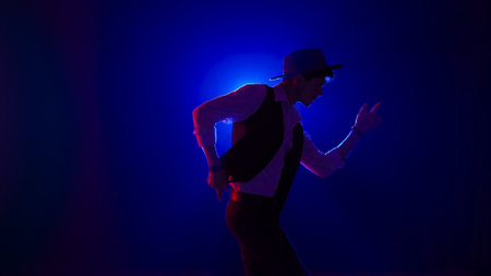 Man musician in stylish hat dancing on dark concert stage against blue spotlight and singing looking at camera, isolated on black studio background.の写真素材