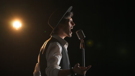 Man musician in stylish hat standing on dark concert stage under spotlight and singing in retro vintage microphone, isolated on black studio background.の写真素材