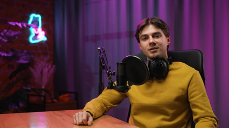 Man host in headphones sitting at table and recording interview video in loft podcast studio using...