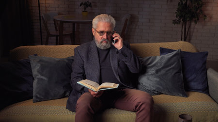 Elder grey haired man spending evening time at home, sitting on the sofa in living room, reading a book and talking on smartphone.. Daily routine lifestyle concept.の写真素材
