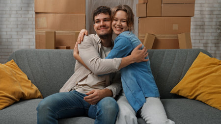 Young couple moving into new apartment, space filled with cardboard boxes, man and woman sit on the sofa in the room of new apartment, hugging positive expression.の写真素材