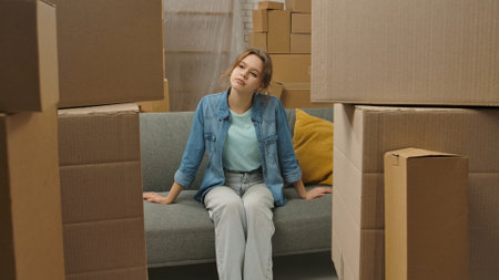 Young couple moving into new apartment, space filled with cardboard boxes, woman sits on the sofa in...