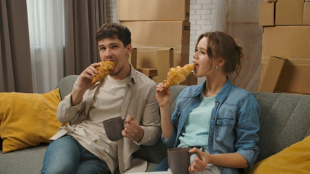 Young couple moving into new apartment, space filled with cardboard boxes, man and woman sit on the sofa look around, eating croissants and drink coffee, positive expression.の写真素材